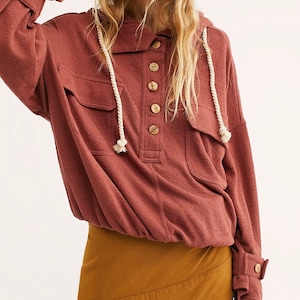 Op de afbeelding: Een roestkleurig hooded shirt met een knoopsluiting aan de voorkant en een grote zak op de borst. Het shirt heeft een capuchon met trekkoord en lange mouwen met knopen aan de manchetten. Het shirt wordt gecombineerd met een bruine rok.