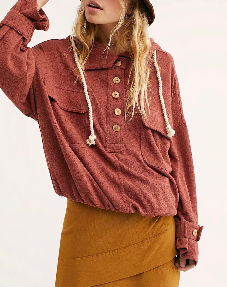 Op de afbeelding: Een roestkleurig hooded shirt met een knoopsluiting aan de voorkant en een grote zak op de borst. Het shirt heeft een capuchon met trekkoord en lange mouwen met knopen aan de manchetten. Het shirt wordt gecombineerd met een bruine rok.