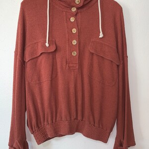 Op de afbeelding: Een roestkleurig hooded sweatshirt met een knoopsluiting aan de voorkant en twee grote zakken. Het sweatshirt heeft een capuchon met trekkoord en elastiek aan de onderkant.