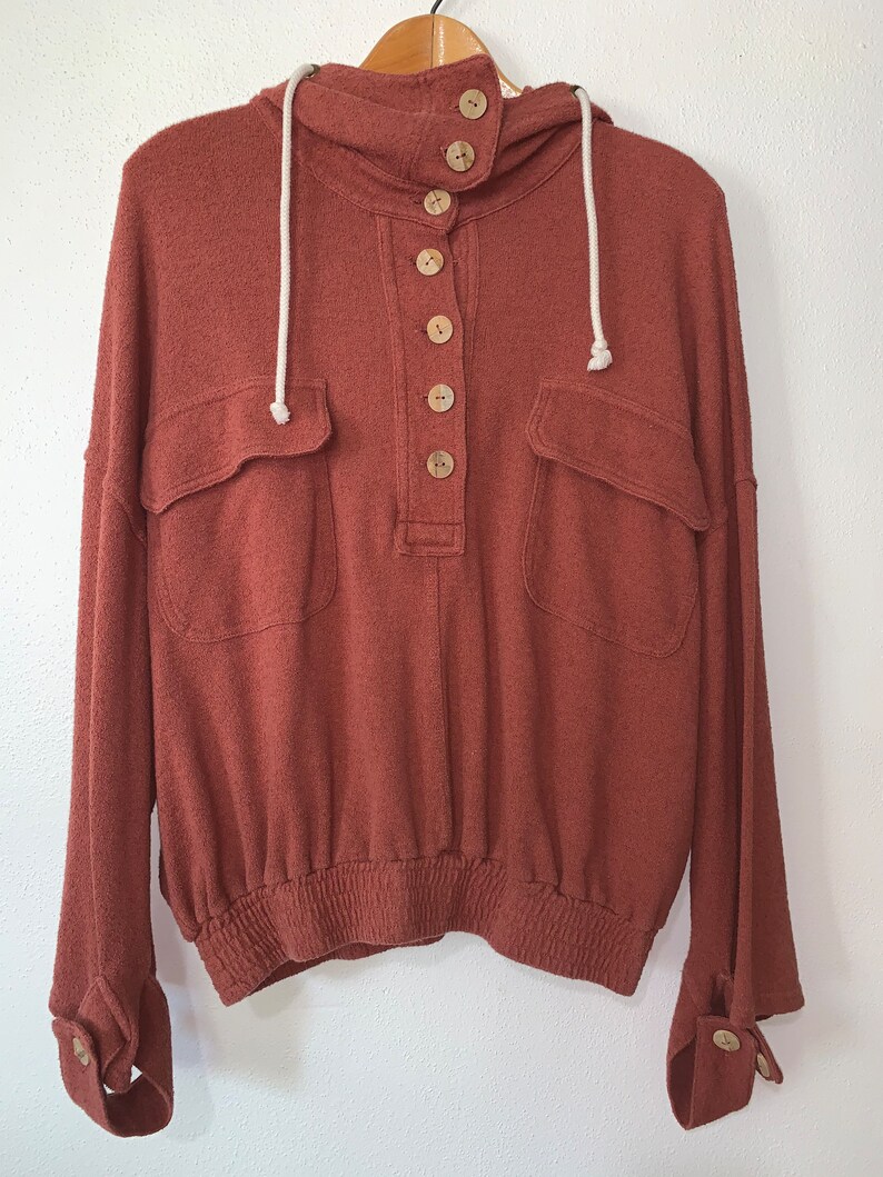 Op de afbeelding: Een roestkleurig hooded sweatshirt met een knoopsluiting aan de voorkant en twee grote zakken. Het sweatshirt heeft een capuchon met trekkoord en elastiek aan de onderkant.