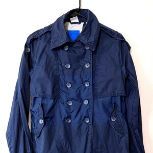 May include: A navy blue double-breasted trench coat with a notched collar and epaulets. The coat features multiple buttons and a classic design, hanging on a black hanger against a white background.
