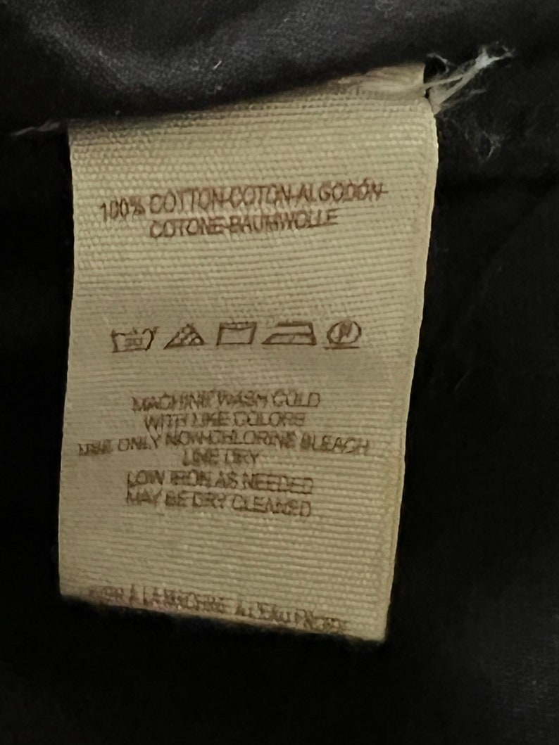 Może przedstawiać: Beige etykieta pielęgnacyjna z tkaniny z instrukcjami prania, suszenia i prasowania. Etykieta zawiera napis "100% Bawełna Coton Algod&oacute;n Cotone Baumwolle", "Prać w pralce w zimnej wodzie z podobnymi kolorami", "Używać tylko wybielacza bez chloru", "Suszyć na linie", "Prasować w niskiej temperaturze w razie potrzeby", "Można czyścić chemicznie".