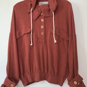 Op de afbeelding: Een roestkleurig hooded sweatshirt met een knoopsluiting, twee klepzakken en witte trekkoordjes. Het sweatshirt heeft een elastische tailleband en gemanchetterd mouwen met een knoop op elke manchet.