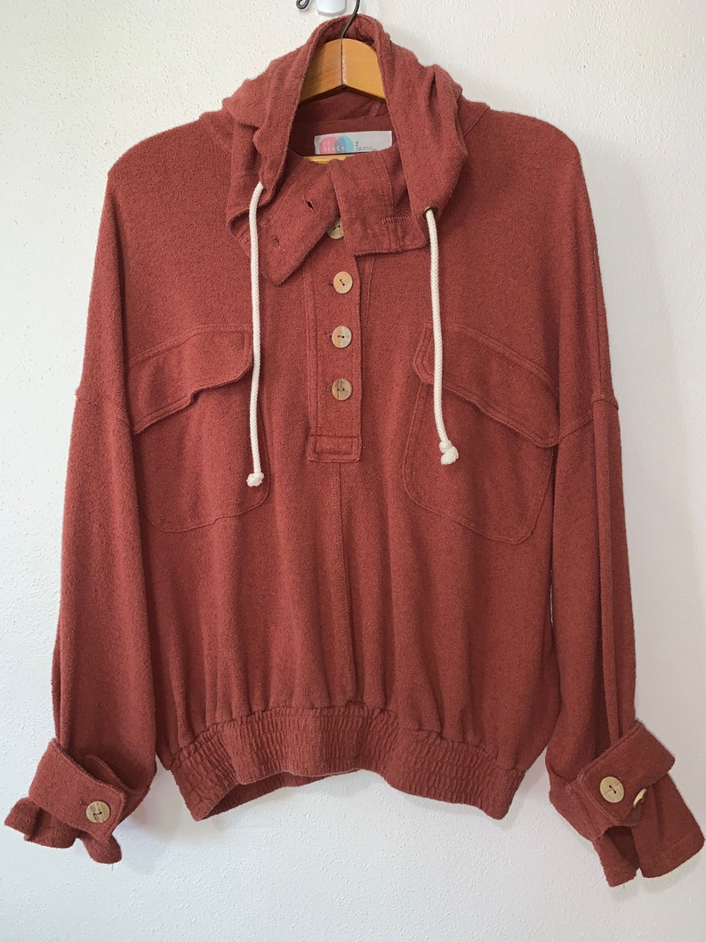 Op de afbeelding: Een roestkleurig hooded sweatshirt met een knoopsluiting, twee klepzakken en witte trekkoordjes. Het sweatshirt heeft een elastische tailleband en gemanchetterd mouwen met een knoop op elke manchet.