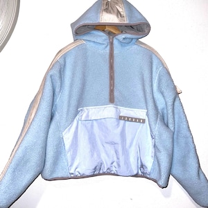 May include: A light blue fleece pullover hoodie featuring a half-zip closure, a large front pouch pocket with the text JORDAN printed on a grey tab, and contrasting beige panels along the shoulders and hood. The garment has a grey trim.