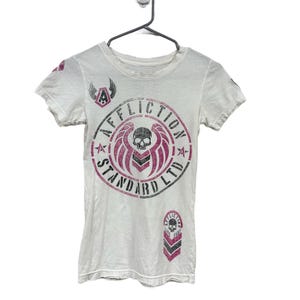 May include: A white short-sleeve t-shirt featuring a graphic print on the front. The design includes a central skull emblem with pink wings, surrounded by the text "AFFLICTION STANDARD LTD" in black. A smaller matching logo appears on the sleeve.