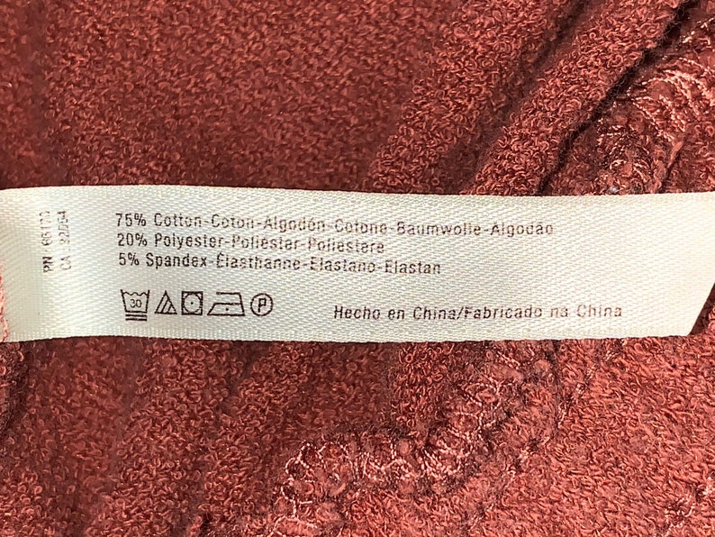 Op de afbeelding: Een wit stoffen label met bruine tekst en wasinstructies. Het label vermeldt: 75% Katoen-Coton-Algod&oacute;n-Cotone-Baumwolle-Algod&atilde;o 20% Polyester-Poliester-Poliestere 5% Spandex-Elasthanne-Elastano-Elastan Hecho en China/Fabricado na China. Het label bevat ook een wasinstructiesymbool.