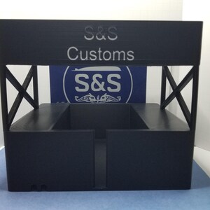 May include: A black display stand with the text "S&S Customs" on the top. The stand has an open rectangular design with support beams and a logo in the background. The stand is made of a solid, dark material.