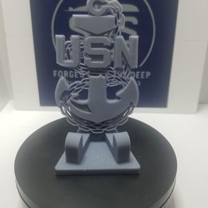 May include: A gray 3D-printed USN anchor emblem. The emblem features the letters "USN" above an anchor design, with a chain wrapping around it. The base has the text "348 Customs". The background has the text "Forged in the Deep".