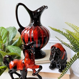 Peut inclure: Ensemble de trois figurines en céramique dans un glacis noir et orange. Les figurines sont un cheval, un chien et un pichet. Le cheval et le chien sont debout sur un livre.