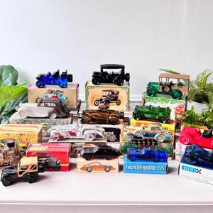May include: A collection of vintage car-shaped bottles in various colors, including blue, green, red, and black. The bottles are displayed on small boxes with vintage car illustrations. Some bottles have the "Avon" brand.