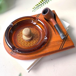 Può includere: Un supporto per fumatori in legno con un posacenere circolare in vetro ambrato, caratterizzato da una base con motivo a diamante e un pomello centrale. Una pipa in legno marrone con bocchino nero poggia in una fessura intagliata.