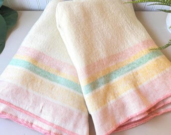May include: A folded cream-colored wool blanket featuring horizontal stripes in pastel pink, yellow, and mint green. The edges of the blanket are finished with a simple pink stitched hem, providing a soft texture and a clean, uniform appearance.