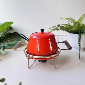 May include: A vintage red fondue pot with a black handle and lid knob. The pot sits on a chrome stand with wooden handles. A classic piece for entertaining, perfect for cheese or chocolate fondue.