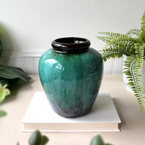 May include: A glossy, green ceramic vase with a dark brown rim. The vase has a rounded body and a smooth, reflective surface. The color transitions from a deep green at the top to a darker shade at the base. The vase is sitting on a white book.