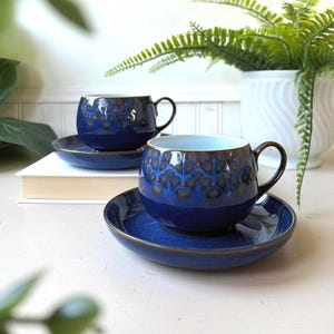May include: Two blue ceramic teacups and saucers. The cups have a dark blue glaze with a pattern of dark brown and red spots. The saucers are a solid blue. The cups have brown handles and a light blue interior.
