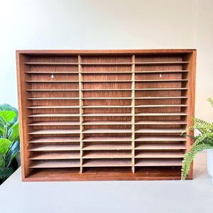 May include: A vintage wooden display shelf with multiple horizontal compartments. The shelf is rectangular with a dark brown frame and light brown shelves. It is designed for organizing and displaying small items.