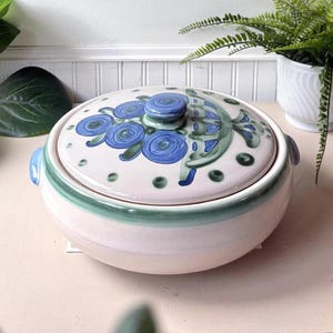 Puede incluir: Una fuente redonda de cerámica para horno con tapa a juego, que presenta una base de color crema con motivos florales azules pintados a mano y detalles en verde. La tapa tiene un pomo central y el cuenco incluye dos asas laterales.