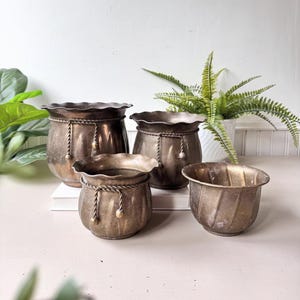 May include: A set of four antique bronze-toned metal planters. The planters have a ruffled rim and decorative rope detailing. The largest planter is approximately 8 inches in diameter, and the smallest is about 4 inches.