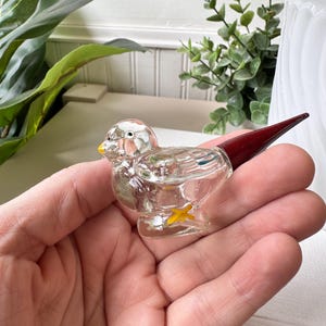 May include: A clear glass bird figurine with a yellow beak and feet, and a dark red tail. The bird is held in a hand, showcasing its delicate details and craftsmanship. The figurine is a decorative item.