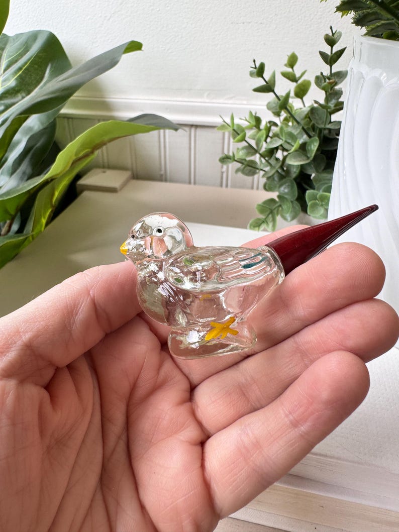 May include: A clear glass bird figurine with a yellow beak and feet, and a dark red tail. The bird is held in a hand, showcasing its delicate details and craftsmanship. The figurine is a decorative item.