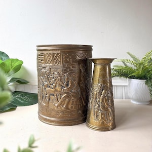 Peut inclure: Un grand seau cylindrique en laiton et un vase conique plus petit en laiton, tous deux ornés de scènes en relief représentant des personnages attablés dans une taverne, avec des détails de briques et de fenêtres sur les surfaces.