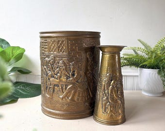 May include: A large cylindrical brass bucket and a smaller tapered brass vase, both featuring embossed relief scenes of figures gathered around a table in a tavern setting with detailed brickwork and window patterns on the exterior surfaces.