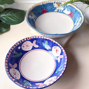 May include: Two round ceramic bowls with scalloped edges. The front bowl features a deep blue interior rim decorated with white fish illustrations, green seaweed accents, and a red dotted border. The back bowl has a blue rim with yellow dots.
