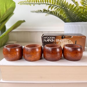 May include: Four brown wooden napkin rings on a book. The box they came in is visible in the background. The box says "Wooden Napkin Rings".