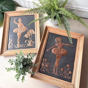 May include: Two framed copper relief art pieces depicting ballerinas in motion. The artwork features a ballerina in a tutu, with floral and plant details. The frames are a light brown color, and the background is a dark gray.