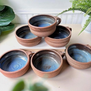 Puede incluir: Un conjunto de cinco cuencos de cerámica redondos con asas, que presentan un exterior texturizado y moteado de color terracota y un interior liso y brillante esmaltado en tonos azul profundo y negro, dispuestos en un grupo.