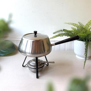 May include: A stainless steel fondue pot with a matching lid and a long black handle. The pot rests on a black metal stand that features a small, circular burner component with a silver handle positioned underneath the main cooking vessel.
