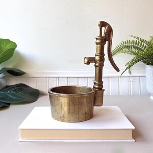 Op de afbeelding: Een koperen waterpompsculptuur met een verticale hendel en een cilindrische bak als basis met verticale lijndetails. Het object is vervaardigd uit een metalen materiaal met een goudkleurige afwerking en een verweerde patina.
