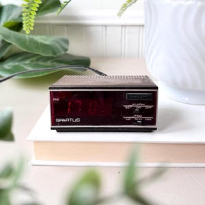 Puede incluir: Un despertador digital Spartus vintage con una parte superior de imitación madera y una pantalla digital roja que muestra las 17:00. El reloj tiene ajustes de alarma y está sobre una pila de libros. Un cable de alimentación negro se extiende desde la parte posterior.