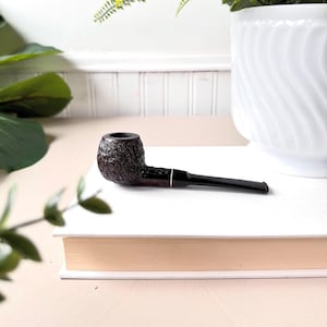 Peut inclure: Une pipe à tabac marron foncé avec un foyer texturé et un tuyau noir. La pipe repose sur un livre blanc, avec un pot décoratif blanc et de la verdure en arrière-plan. La pipe a un design classique et traditionnel.