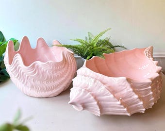 May include: Two pale pink ceramic bowls shaped like large seashells. The bowls feature intricate, wavy, and ridged textures on their exterior surfaces, mimicking the natural form of giant clams and conch shells with a smooth, glossy finish.