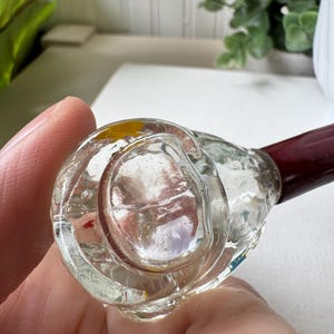 May include: Clear glass pipe with a dark red stem. The bowl of the pipe is round and has a textured surface. The pipe is held in a person's hand.