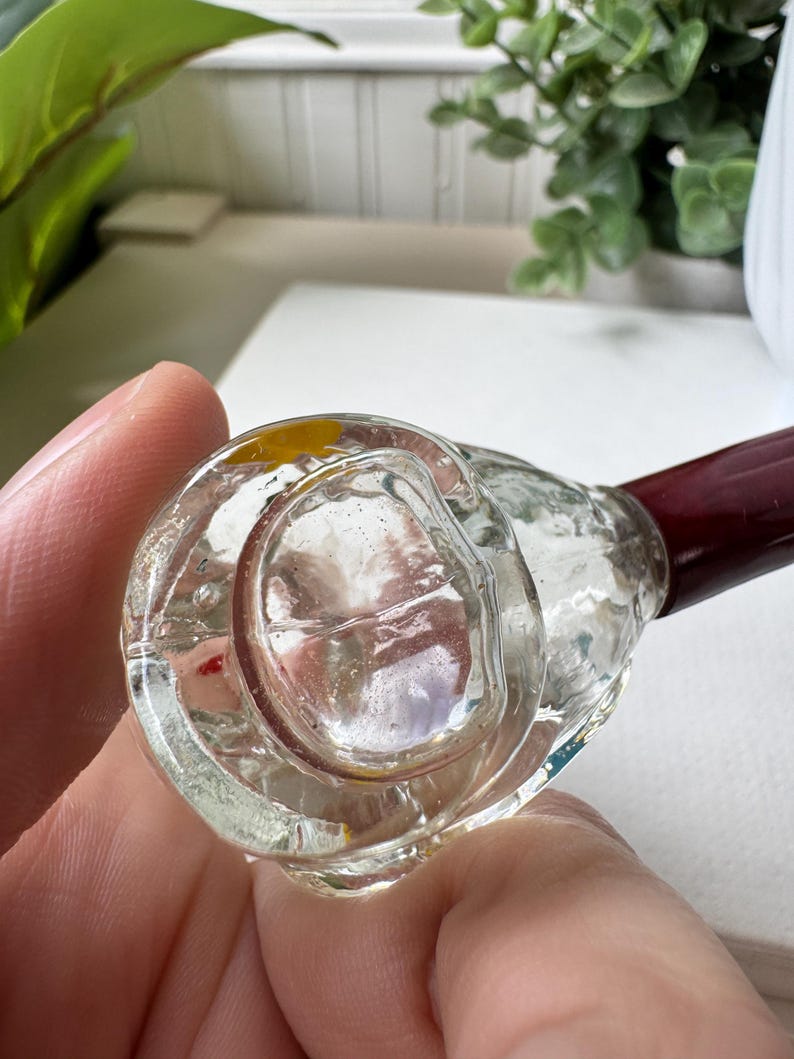 May include: Clear glass pipe with a dark red stem. The bowl of the pipe is round and has a textured surface. The pipe is held in a person's hand.