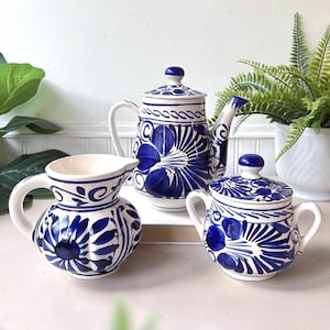 Könnte beinhalten: Ein Keramik-Teeservice bestehend aus einer Teekanne mit Deckel, einem Milchkännchen und einer Zuckerdose mit Deckel. Jedes Teil hat eine weiße Basis mit handgemalten, tiefblauen Blumen- und Wirbelmustern, inklusive zentraler Blüte.