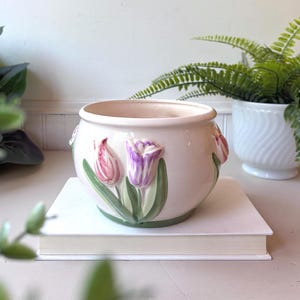 May include: A round, cream-coloured ceramic planter featuring embossed, hand-painted tulip flowers in shades of pink and purple with green stems and leaves. The planter has a smooth, glossy finish and a wide, open top, showcasing a floral design.