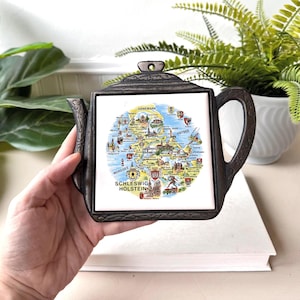 May include: A dark metal trivet in the shape of a teapot, featuring a map of Schleswig-Holstein on a white tile. The map is illustrated with landmarks and the text "Schleswig Holstein". The trivet has a handle and spout, ideal for protecting surfaces from heat.