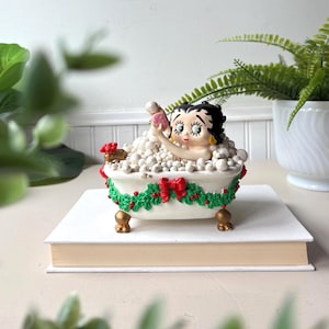 May include: A figurine of a cartoon character with black hair and large eyes sitting in a white clawfoot bathtub filled with white bubbles. The tub is decorated with a green garland, red berries, and a red bow. It has gold-colored feet.