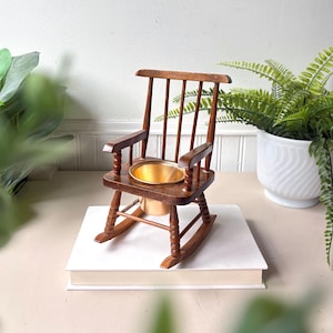 May include: Miniature wooden rocking chair with a brass pot. The chair is a dark brown colour with a classic rocking chair design. The brass pot is circular and sits in the seat of the chair. The chair is placed on top of a stack of white books.