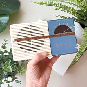 May include: A vintage Sony radio in cream and blue. The radio features a speaker grill, a tuning dial, and the brand name "SONY". The rectangular radio appears to be well-preserved, showcasing a retro aesthetic.