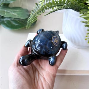 以下が含まれることがあります： 光沢のある濃い青色の釉薬を施したセラミック製のカメの置物。カメは詳細な甲羅とヒレを持ち、小さなステッカーが背中に貼られています。この置物は装飾品です。