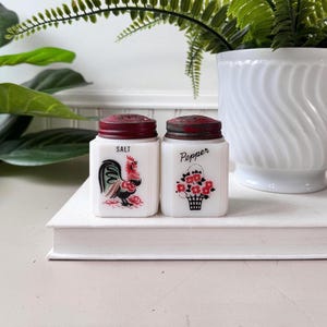 Puede incluir: Un juego de salero y pimentero vintage. Los saleros de vidrio blanco tienen tapas de metal burdeos. Uno presenta una ilustración de gallo, el otro una cesta floral. Las palabras "Salt" y "Pepper" están impresas.