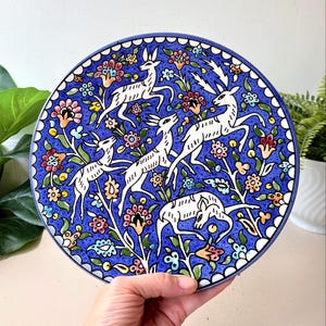 May include: A round ceramic decorative plate featuring a vibrant blue background with a detailed hand-painted pattern of four white deer leaping among colorful stylized flowers, leaves, and vines, finished with a scalloped blue and white border.