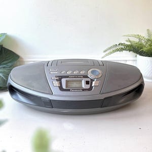 Puede incluir: Un sistema estéreo portátil Panasonic Power Blaster de color gris con una platina de casete de carga superior, una pantalla digital central que muestra TAPE 909, altavoces integrados y varios botones de control para reproducción y volumen.