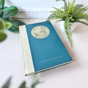 Op de afbeelding: Een teal hardcover boek getiteld "The Reader's Digest Great World Atlas". Het boek heeft een gouden wereldkaart op de omslag en een crèmekleurige rug. Het boek ligt op een lichtroze oppervlak.