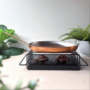 Puede incluir: Una sartén redonda de cobre con un mango largo de latón sobre un soporte calentador de metal negro. El soporte cuenta con dos elementos calefactores circulares y una base rectangular. La sartén tiene una superficie interior plateada.